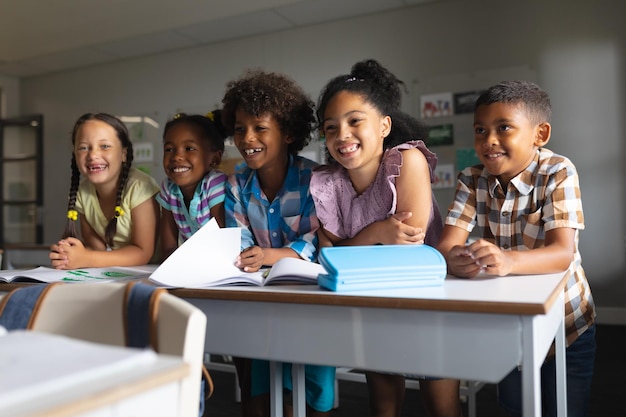 Photo des élèves d'école primaire multiraciaux heureux assis à leur bureau en classe.