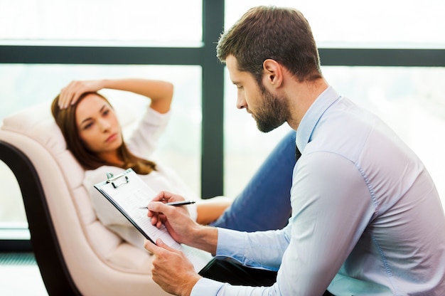 Elle a besoin d'un avis d'expert. Jeune femme frustrée assise à la chaise et tenant la main sur la tête tandis que le jeune homme assis près d'elle et écrivant quelque chose dans son presse-papiers