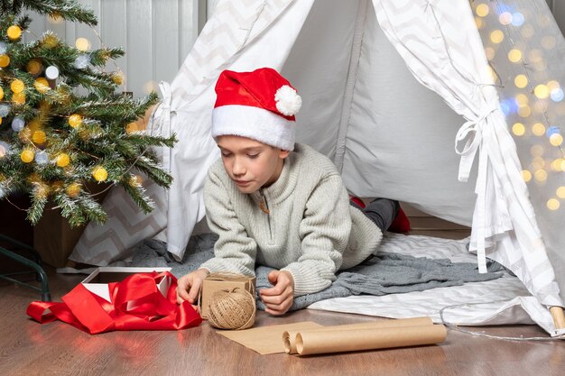 Emballage cadeau Un enfant dans un bonnet rouge enveloppe les cadeaux de Noël surprises dans du papier kraft avec une corde de rubans rouges