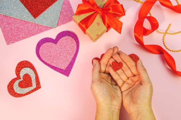 Emballage des cadeaux de Saint Valentin ou d'anniversaire. Cadeaux de la Saint-Valentin avec un coeur en papier rouge sur une surface rose. Vue de dessus. Les mains féminines tiennent le coeur de papier rouge.