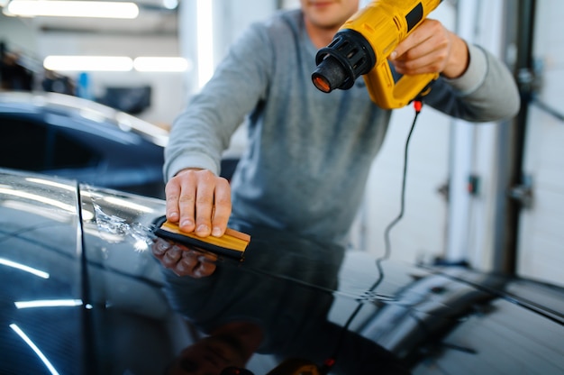 L'emballage masculin contient une raclette et un pistolet thermique, un service de teinture de voiture. Travailleur appliquant une teinte de vinyle sur la fenêtre du véhicule dans le garage, le verre automobile teinté