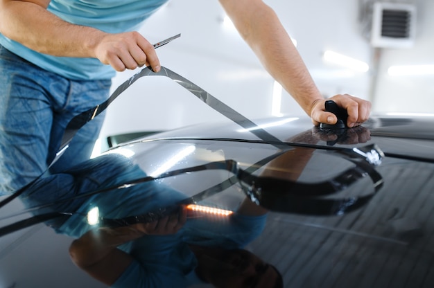 L'emballage masculin coupe la teinte de la voiture, le service de réglage. Travailleur appliquant une teinte de vinyle sur la fenêtre du véhicule dans le garage, le verre automobile teinté