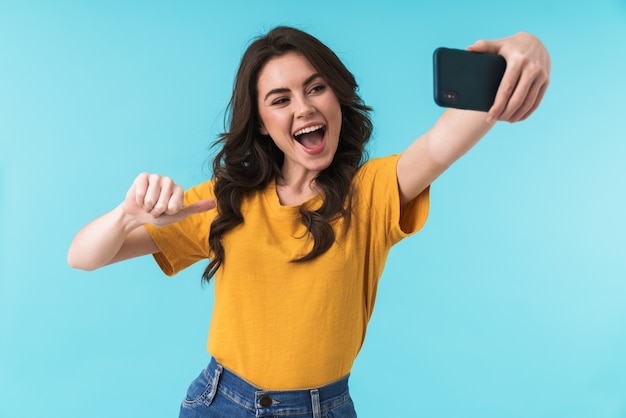 émotif heureux optimiste jeune jolie femme posant isolé sur mur bleu prendre selfie par téléphone mobile pointant sur elle-même.