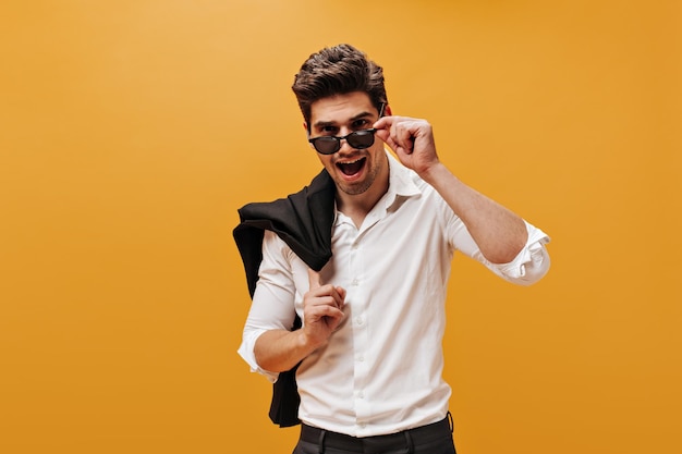 Émotionnel jeune homme brune en chemise blanche élégante mettant des lunettes de soleil et tenant une veste noire sur fond isolé orange