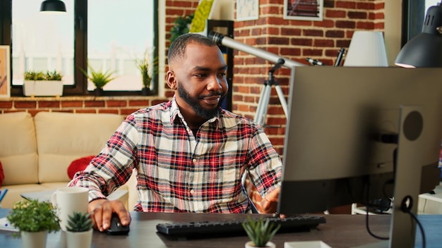 Employé afro-américain fatigué et endormi travaillant à distance, tapant des données sur un ordinateur, heureux de terminer bientôt son travail. Télétravailleur répondant aux e-mails d'emploi dans un appartement chaleureux et confortable