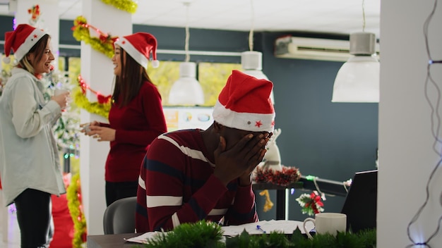 Employé afro-américain travaillant dans un bureau décoré de Noël, essayant de se concentrer pendant que des collègues insouciants profitent d'une pause-café ensemble. Travailleur avec un chapeau de Père Noël frustré par des collègues distrayants