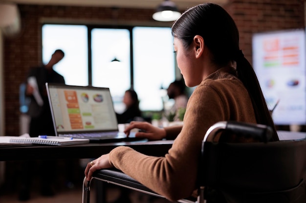 Employé asiatique handicapé physique assis en fauteuil roulant, travaillant sur un ordinateur portable dans un bureau adapté aux personnes handicapées. Travailleuse souffrant d'un problème de santé chronique analysant la présentation du site Web.