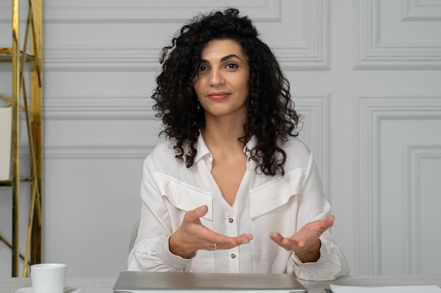 Employé aux cheveux bouclés souriant et satisfait assis à un bureau dans le bureau en regardant la caméra un