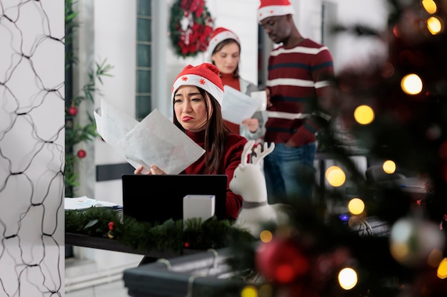 Employé bourreau de travail déprimé contrarié par des documents stressants de l'entreprise en retard à Noël. Employé stressé se surmenant dans un lieu de travail décoré de façon festive, essayant de terminer un projet difficile