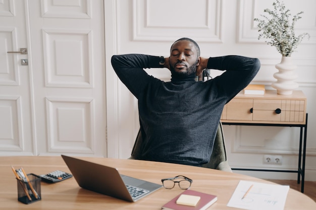 Employé de bureau afro-américain surmené fatigué se reposant les yeux fermés et les mains derrière la tête