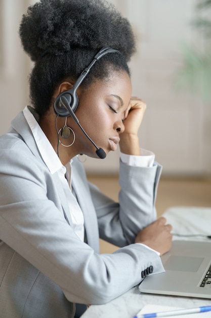 Un employé de bureau afro fatigué avec des écouteurs dort sur le lieu de travail, souffrant de fatigue chronique