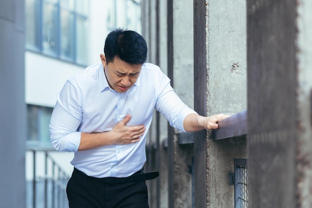 Employé de bureau asiatique malade ayant de graves douleurs à la poitrine homme d'affaires tenant la main sur le cœur à l'extérieur du bureau