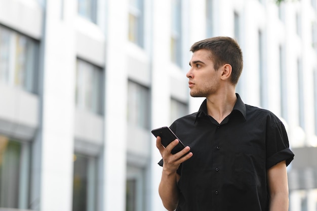 un employé de bureau en chemise noire se tient sur un fond urbain clair avec un smartphone à la main