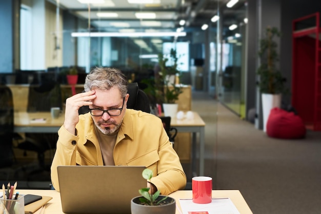 Employé de bureau mature sérieux regardant à travers des données en ligne sur l'écran d'un ordinateur portable