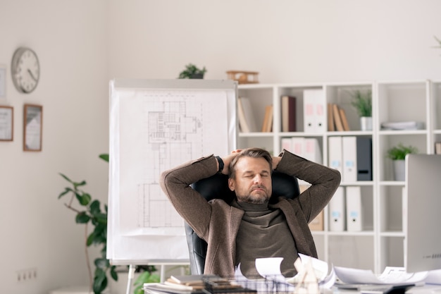 Photo employé de bureau mature en tenues de soirée assis dans un fauteuil par bureau tout en se concentrant sur le travail ou se détendre à la pause