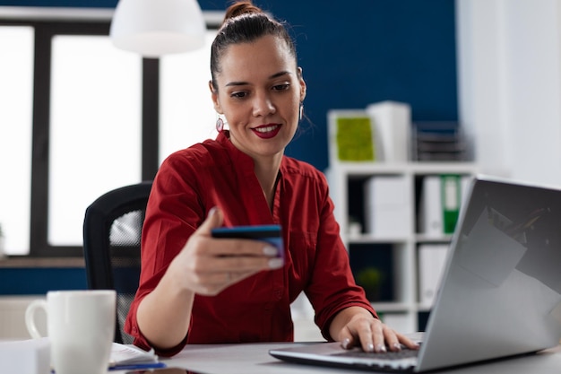 Employé en chemise rouge vérifiant les détails de la carte de crédit pour les achats en ligne. Femme d'affaires au bureau de démarrage utilisant un ordinateur portable pour les services bancaires par Internet. Entrepreneur tapant le numéro de compte assis au bureau.