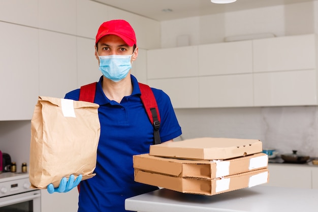 Employé du livreur en bonnet rouge t-shirt gants de masque uniformes donner des boîtes à pizza de commande de nourriture isolées sur fond jaune studio. Service de quarantaine pandémique coronavirus virus grippe 2019-ncov concept