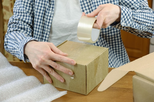 Photo l'employé de l'entrepôt enveloppe la commande de la boutique en ligne avec du scotch dans une boîte en carton. petite entreprise, traitement des commandes et emballage