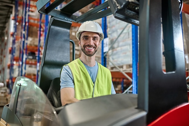 Employé d'entrepôt joyeux souriant dans un casque et un gilet réfléchissant assis dans le chariot élévateur électrique