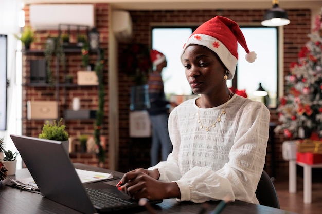 Employé de l'entreprise utilisant un ordinateur portable au travail de bureau, célébrant la saison d'hiver avec des décorations de noël et un arbre. Femme avec bonnet de noel travaillant dans l'espace avec des ornements saisonniers festifs.