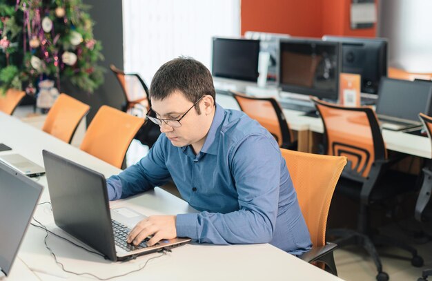 Employé de l'homme en chemise bleue et lunettes assis au bureau au bureau à l'aide d'un ordinateur portable