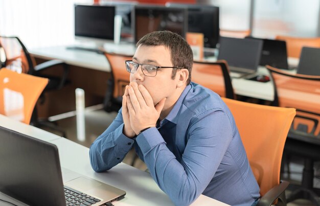 Employé de l'homme en chemise bleue et lunettes assis au bureau au bureau avec un ordinateur portable cachant le visage se sentant bouleversé par la date limite Concept de journée de travail chargée d'affaires Soins de santé contre le stress au travail