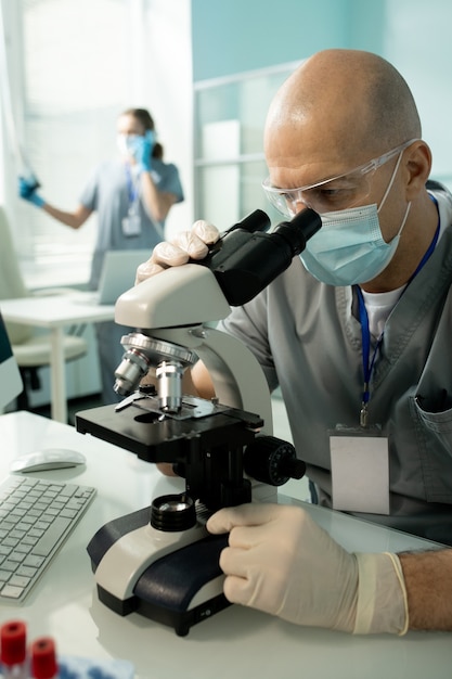 Employé de laboratoire chauve concentré dans des lunettes et des gants en latex assis à table et travaillant avec un microscope tout en faisant des recherches