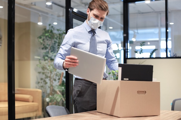Employé de licenciement en masque médical préventif dans un coronavirus épidémique. Un travailleur licencié triste emporte ses fournitures de bureau avec lui.