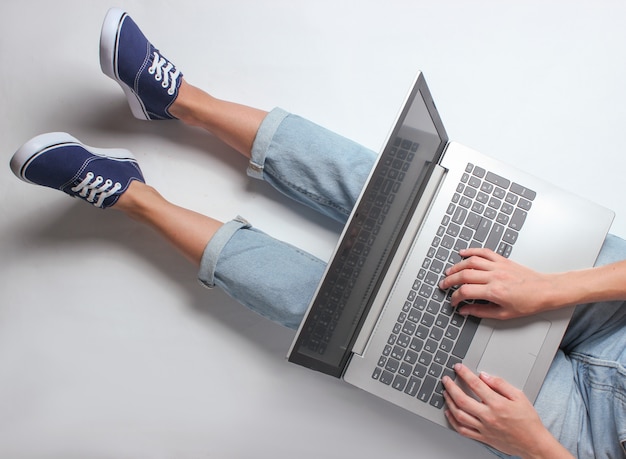 Employé en ligne sur le lieu de travail. Fragment de jambes féminines en jeans et baskets. Une femme est assise et utilise un ordinateur portable. Concept indépendant. Travail à la maison.