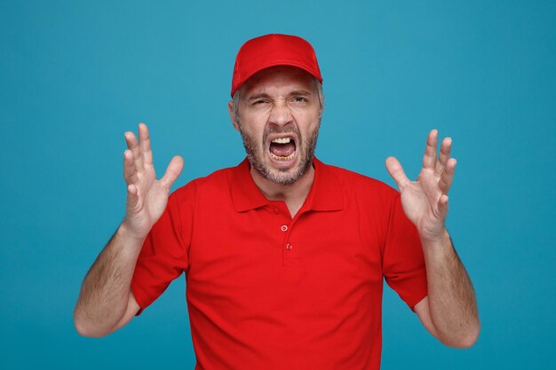 Employé de livreur en casquette rouge uniforme de t-shirt blanc en colère et frustré criant avec les bras levés debout sur fond bleu