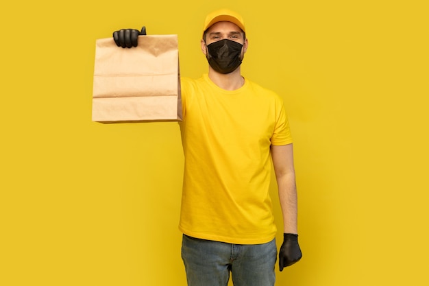 Employé de livreur en chapeau jaune tshirt masque uniforme gant tenir le paquet de papier craft avec de la nourriture isolé