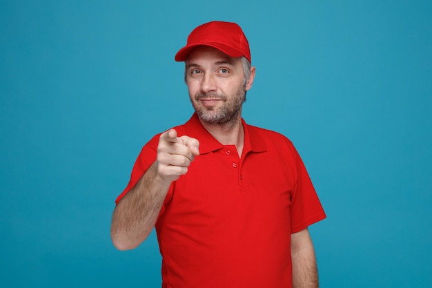 Employé de livreur en uniforme de t-shirt blanc à capuchon rouge regardant la caméra heureux et positif pointant avec l'index à la caméra souriant debout sur fond bleu