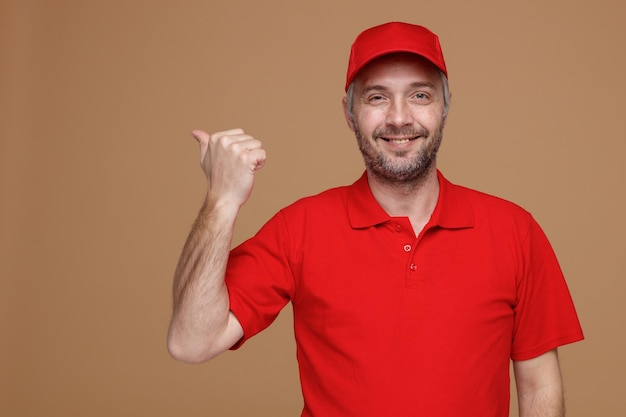 Employé de livreur en uniforme de t-shirt blanc à capuchon rouge regardant la caméra heureux et positif pointant avec le pouce sur le côté souriant debout sur fond marron