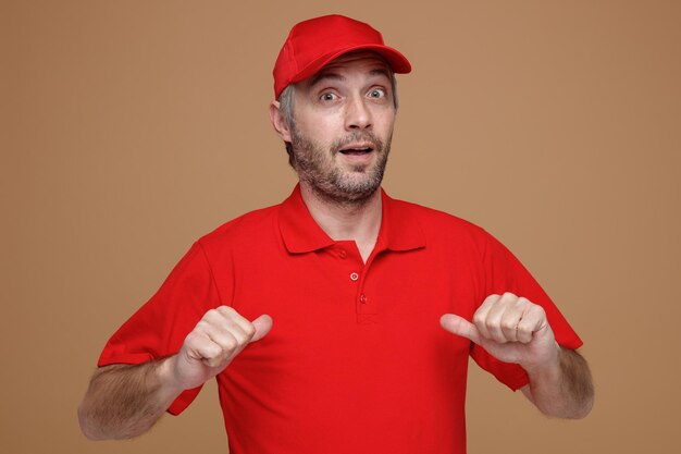 Employé de livreur en uniforme de t-shirt blanc à capuchon rouge regardant la caméra surpris en se montrant debout sur fond marron