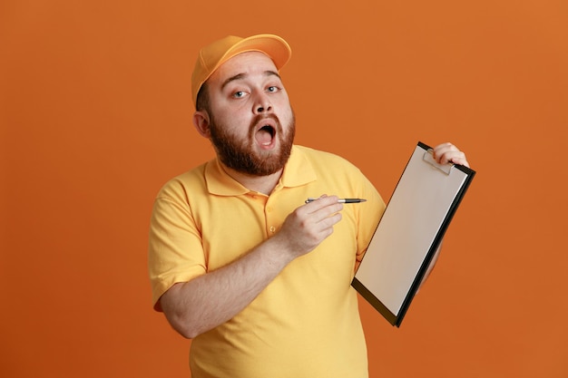 Employé de livreur en uniforme de t-shirt blanc à casquette jaune tenant un presse-papiers et un stylo regardant la caméra étonné et surpris debout sur fond orange