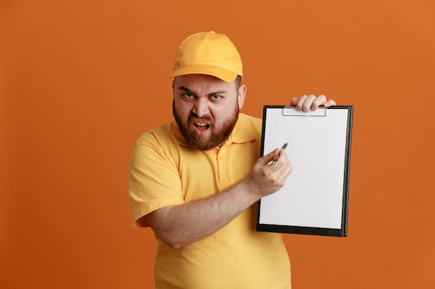 Employé de livreur en uniforme de t-shirt blanc à casquette jaune tenant un presse-papiers et un stylo regardant la caméra avec un visage en colère fronçant les sourcils debout sur fond orange