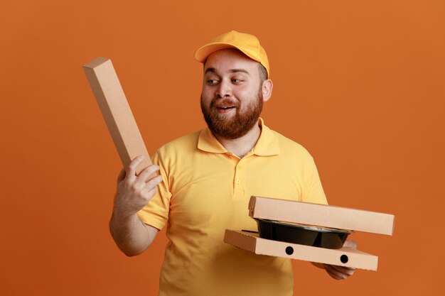 Employé de livreur en uniforme de t-shirt blanc à casquette jaune tenant un récipient de nourriture et des boîtes à pizza regardant la boîte avec le sourire sur le visage debout sur fond orange