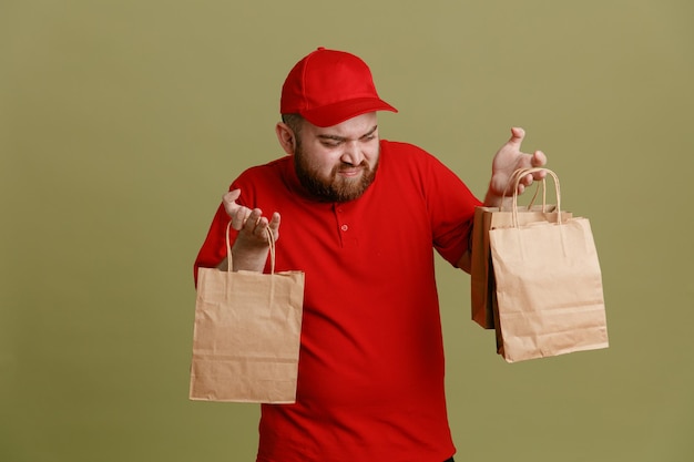 Employé de livreur en uniforme de t-shirt blanc à casquette rouge tenant des sacs en papier en les regardant avec une expression dégoûtée debout sur fond vert