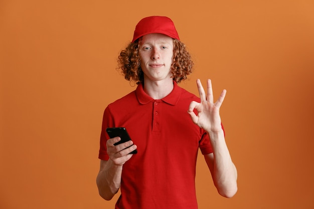 Employé de livreur en uniforme de t-shirt blanc à casquette rouge tenant un smartphone regardant la caméra souriant confiant faisant signe ok debout sur fond orange
