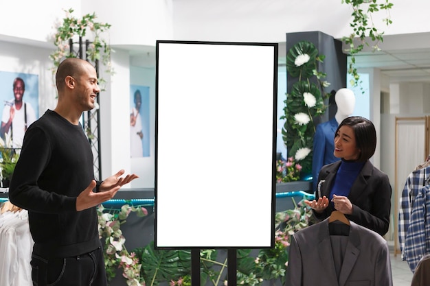 Employé de magasin de vêtements et client discutant de la collection de marques de vêtements tout en se tenant près de la maquette du tableau blanc des publicités. Consultant en boutique utilisant un affichage numérique pour montrer les options de vêtements au client