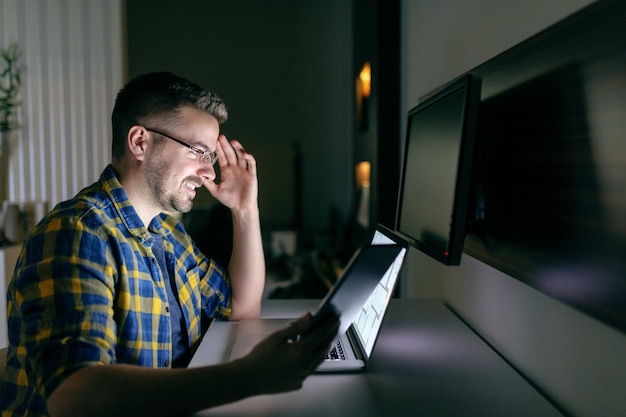 Photo employé mal rasé souriant travaillant avec des lunettes et en chemise à carreaux tenant dans la main tablette tout en regardant un ordinateur portable