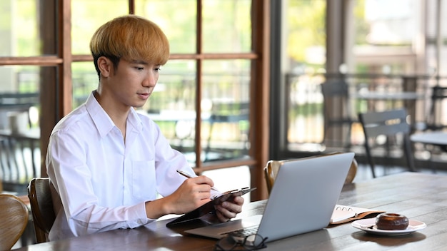 Employé masculin lisant des informations sur un ordinateur portable et prenant des notes sur un ordinateur portable