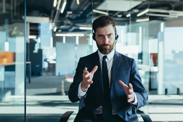 L'employé passe un appel vidéo regarde la caméra et explique la tâche des gestes sérieux avec ses mains travaille dans un bureau moderne utilise un casque