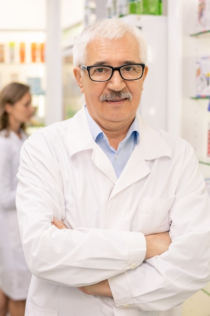 Employé de pharmacie âgé en blouse blanche et lunettes croisant les bras par la poitrine en se tenant debout contre des présentoirs avec des produits médicaux et son collègue