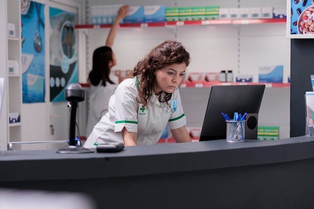 Employé de pharmacie vérifiant la liste des médicaments sur un écran d'ordinateur, analysant le rapport de traitement pharmaceutique pour le client. Femme pharmacienne portant un uniforme médical travaillant dans une pharmacie, soutien médical