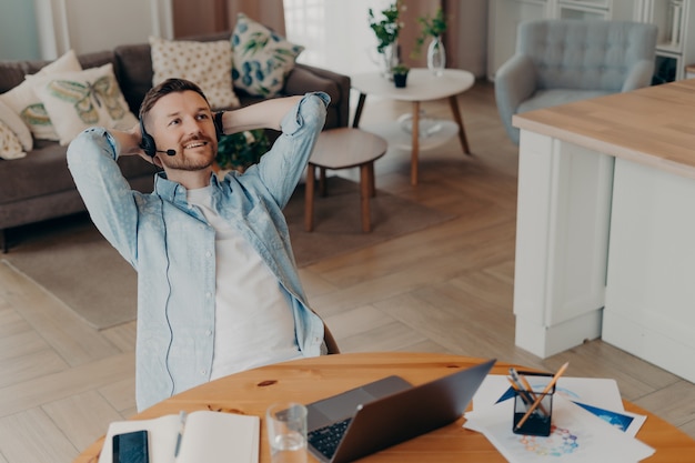 L'employé de sexe masculin se repose après une dure journée de travail garde les mains derrière la tête pense à quelque chose et planifie est assis au bureau avec un bloc-notes pour ordinateur portable écoute le cours d'entreprise audio via un casque fonctionne à distance