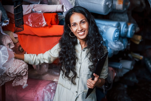 Un employé souriant positif avec un bloc-notes est dans le stockage plein de tissu.