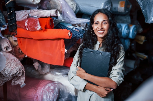 Un employé souriant positif avec un bloc-notes est dans le stockage plein de tissu.