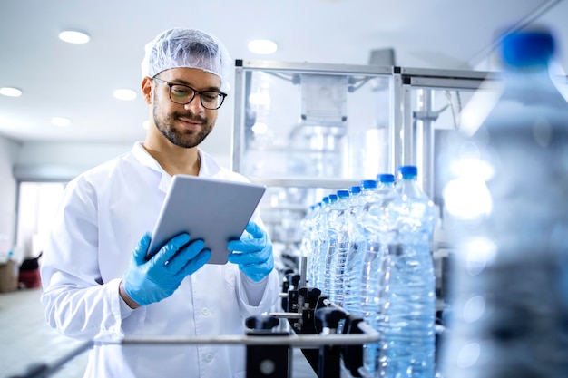 Employé d'usine d'embouteillage ou debout près d'une machine automatisée et vérifiant la production d'eau