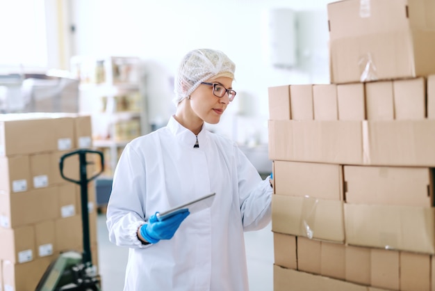 Employée blonde de race blanche en uniforme stérile debout et s'occupant de la logistique des produits. Intérieur de l'usine alimentaire. Tablette en main.